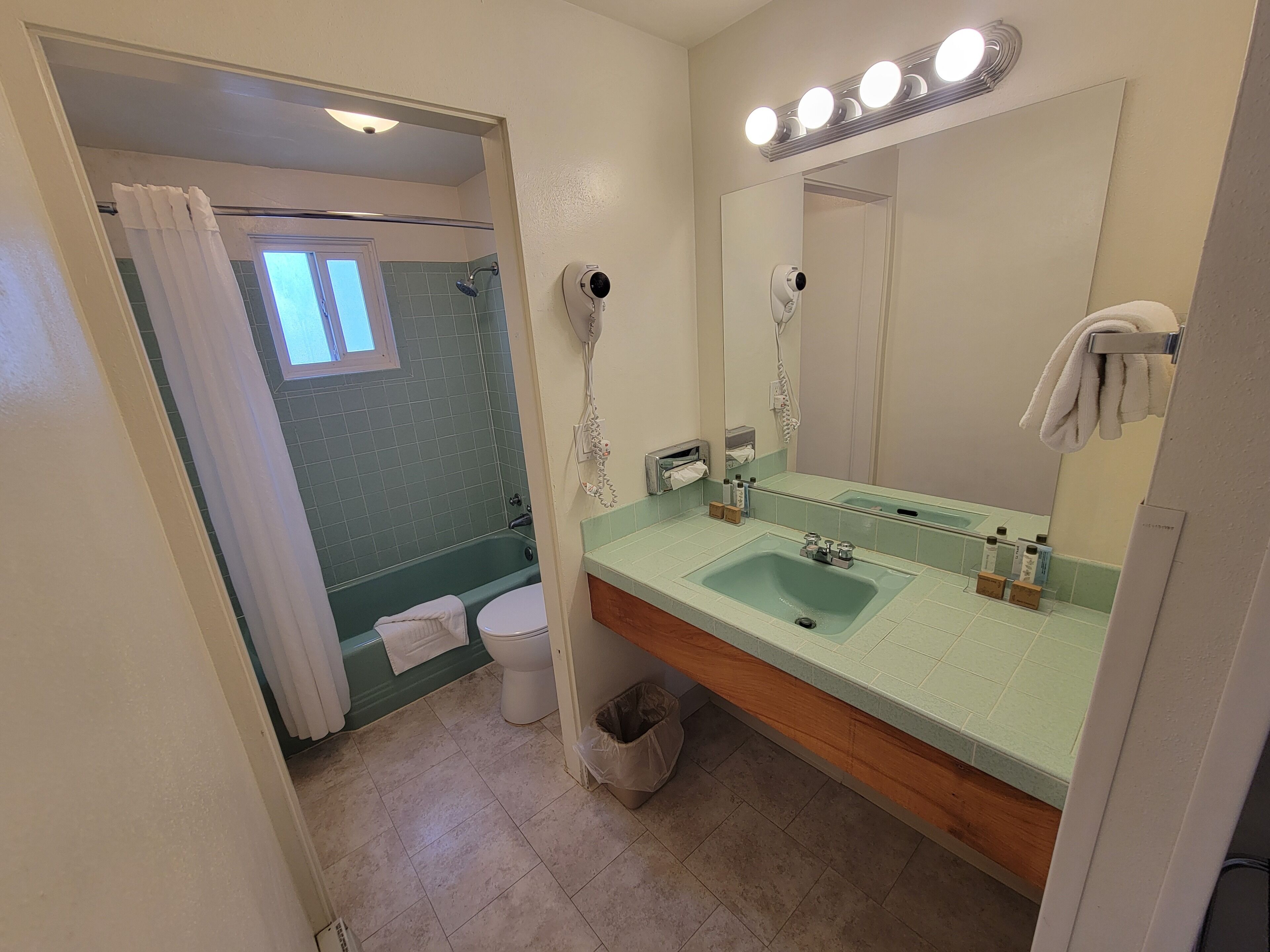 A bathroom with a green-tiled shower, a white toilet, a sink with a mirror above it, and a towel hanging on the wall.