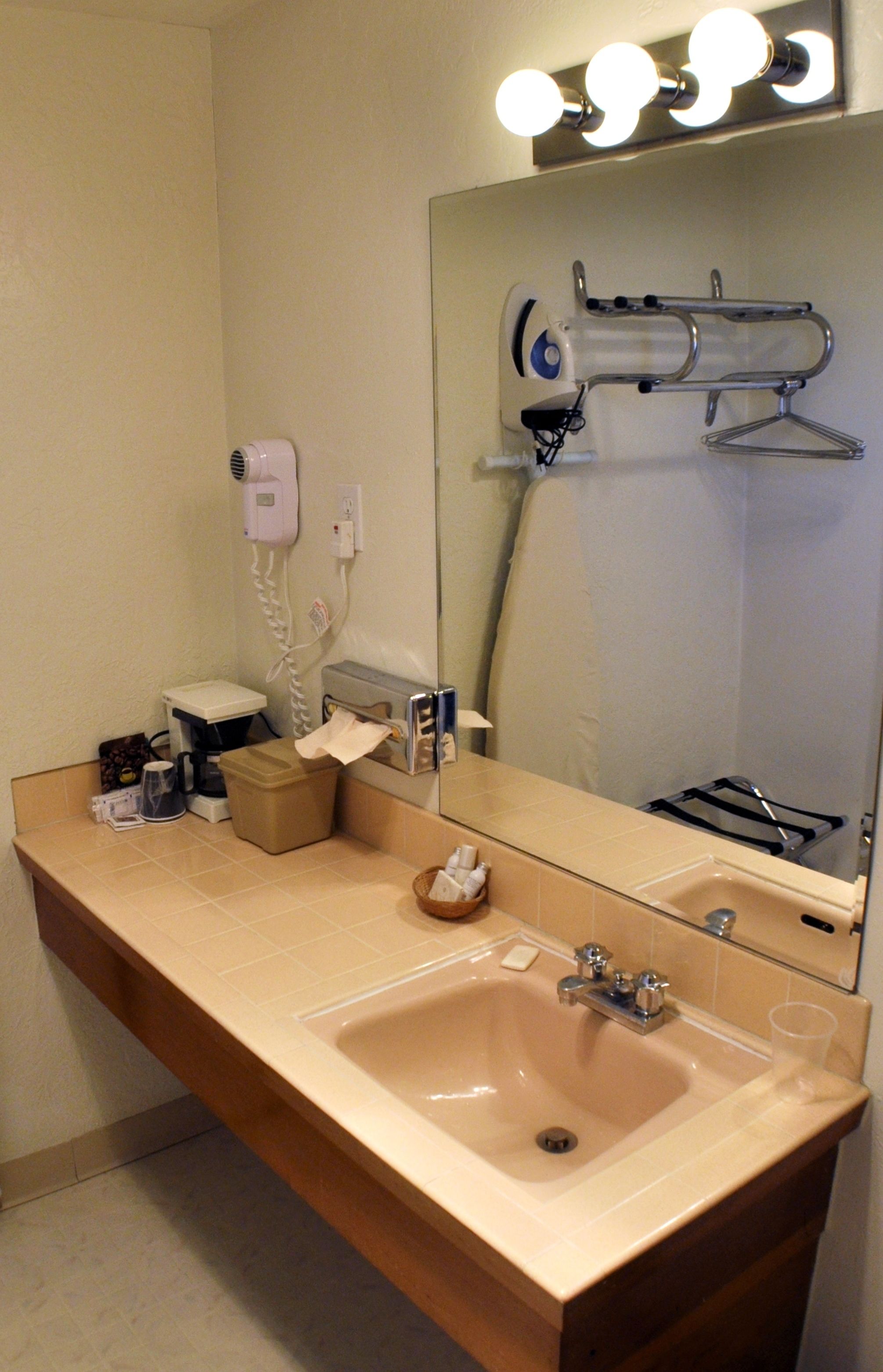 A bathroom with a sink, mirror, and vanity. There is a hair dryer, a towel dispenser, and a soap dish.