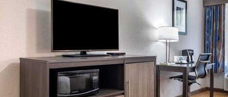Habitación de hotel con televisión de pantalla plana sobre un mueble de madera, microondas, escritorio con silla y lámpara.