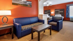 A hotel room with a blue sofa, a small table, a desk with a lamp, a bed with a headboard, and a painting on the wall.
