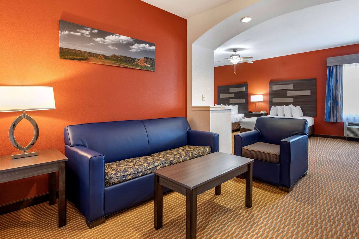 Une chambre d’hôtel avec un canapé bleu, une petite table, un bureau avec une lampe, un lit avec une tête de lit et un tableau au mur.