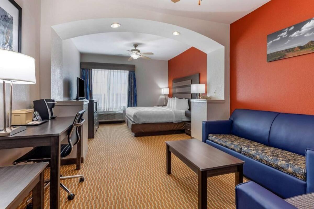Une chambre d’hôtel avec un lit, un bureau, un canapé bleu, un ventilateur de plafond et une grande fenêtre avec des rideaux.