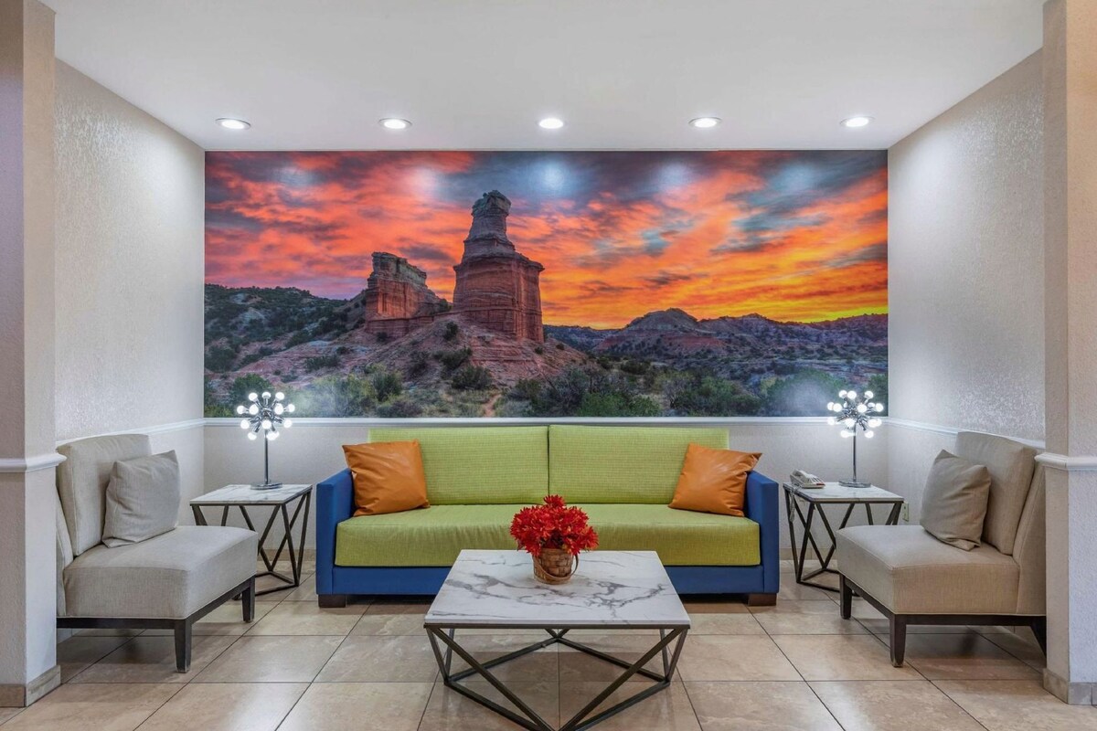 Een kamer met een groene bank, twee fauteuils, een marmeren salontafel en een groot wanddecor van een zonsondergang boven rotsformaties.