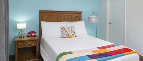 A neatly made bed with a rainbow-colored blanket, a wooden nightstand with a lamp, and a vintage telephone.