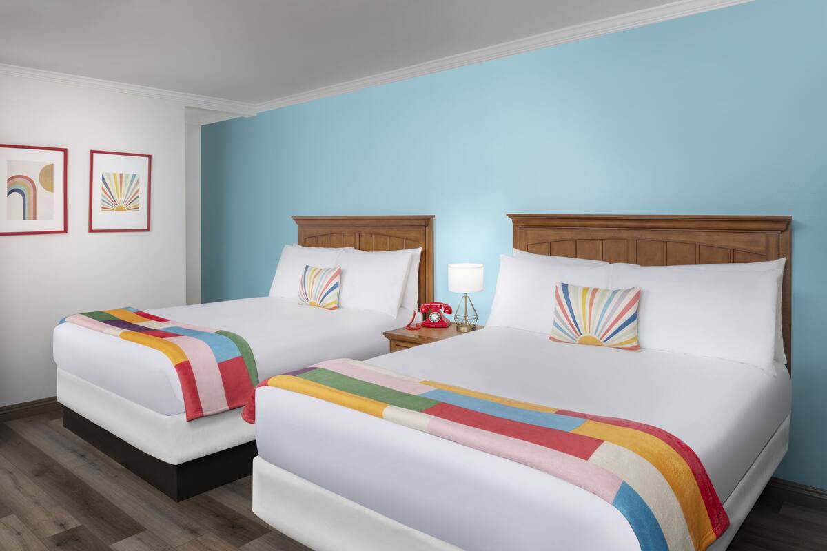 Ein Hotelzimmer mit zwei Betten, jedes mit einer bunten, gestreiften Decke und passenden Kissen. Ein Nachttisch mit einem roten Telefon und einer Lampe steht bereit. Die Wände sind hellblau gestrichen, und es gibt zwei gerahmte Bilder an der Wand.