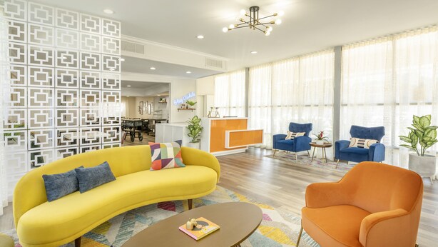 A modern living room with a yellow sofa, an orange armchair, and a coffee table. There is a patterned rug, a white decorative wall, and large windows with sheer curtains. - The Sojourn Los Angeles - Sherman Oaks (Sherman Oaks)