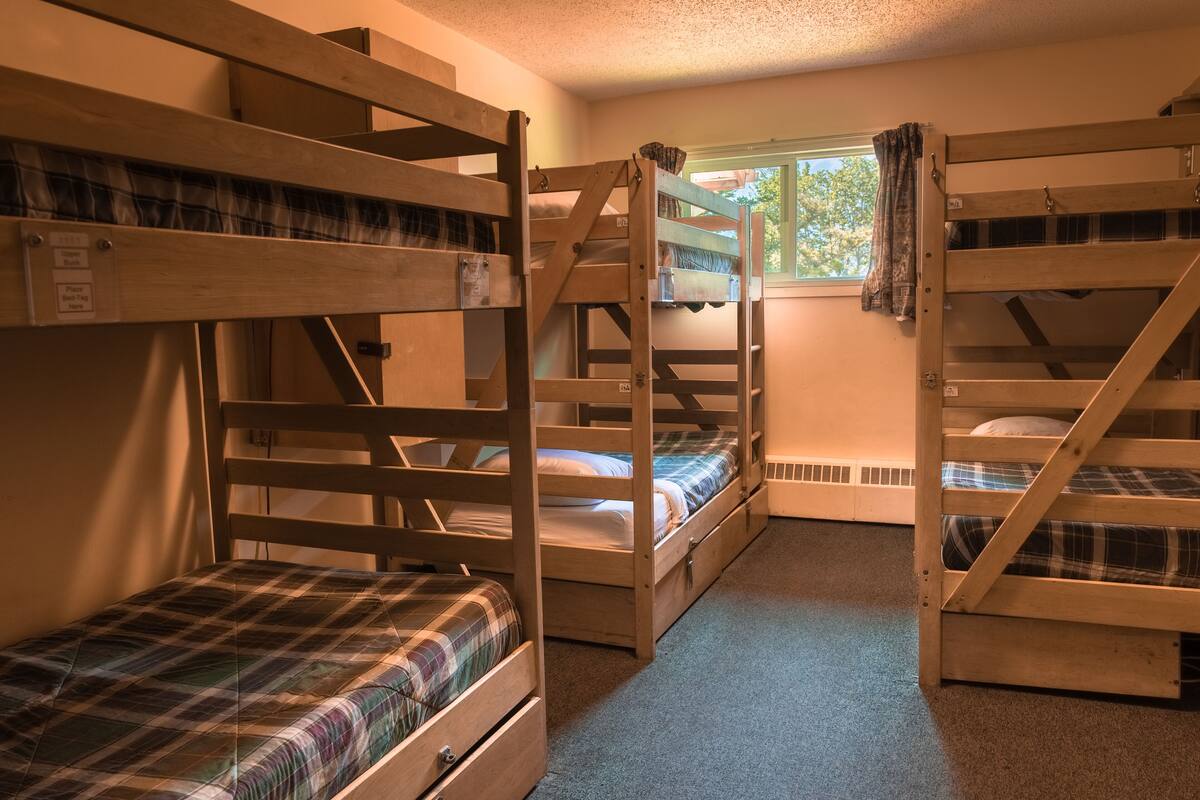 Une chambre avec trois lits superposés, chacun recouvert d’une couverture à carreaux. Il y a une fenêtre d’où l’on peut voir les arbres.