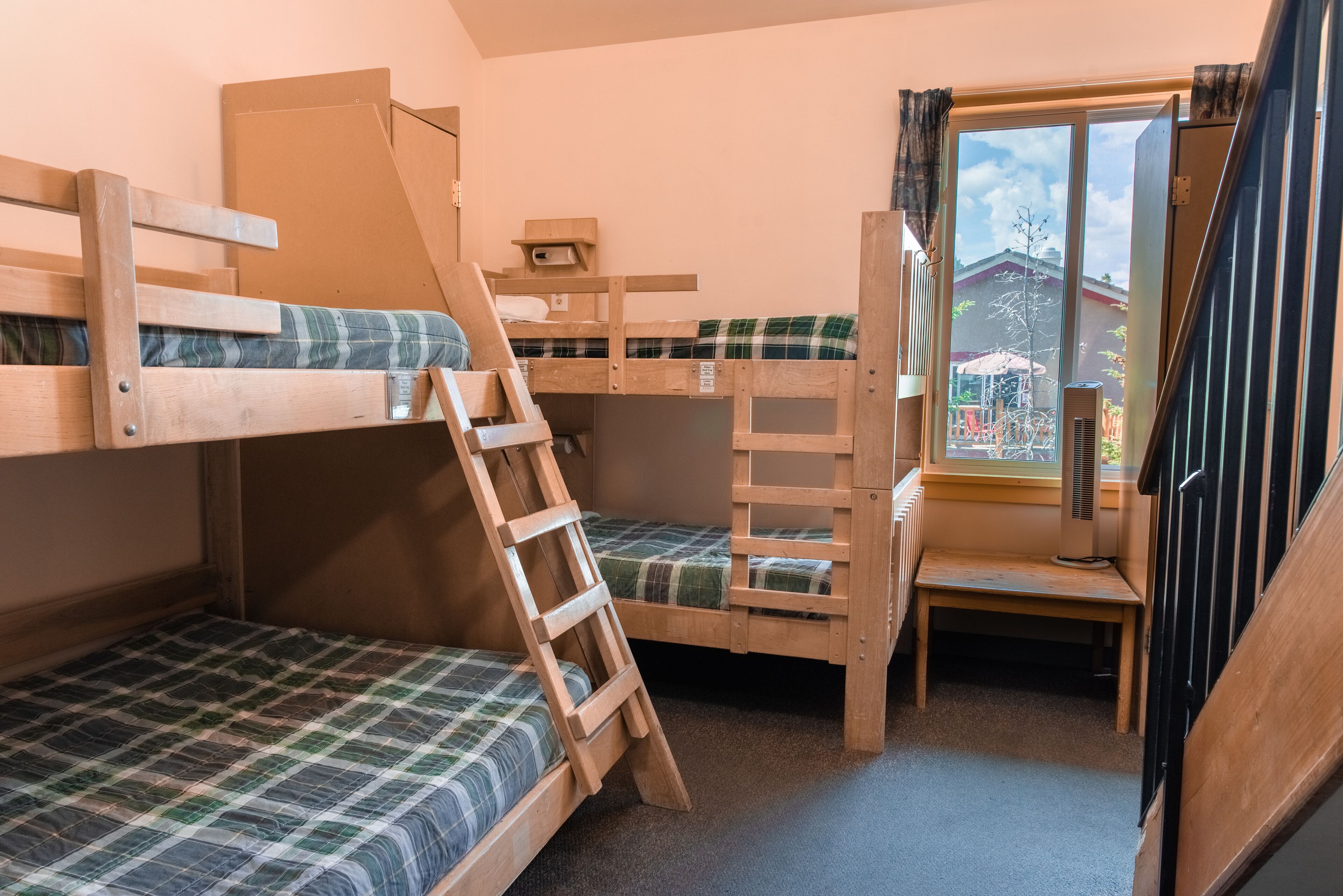 A dormitory room with bunk beds, a ladder, a small table, and a window with a view of the outside.