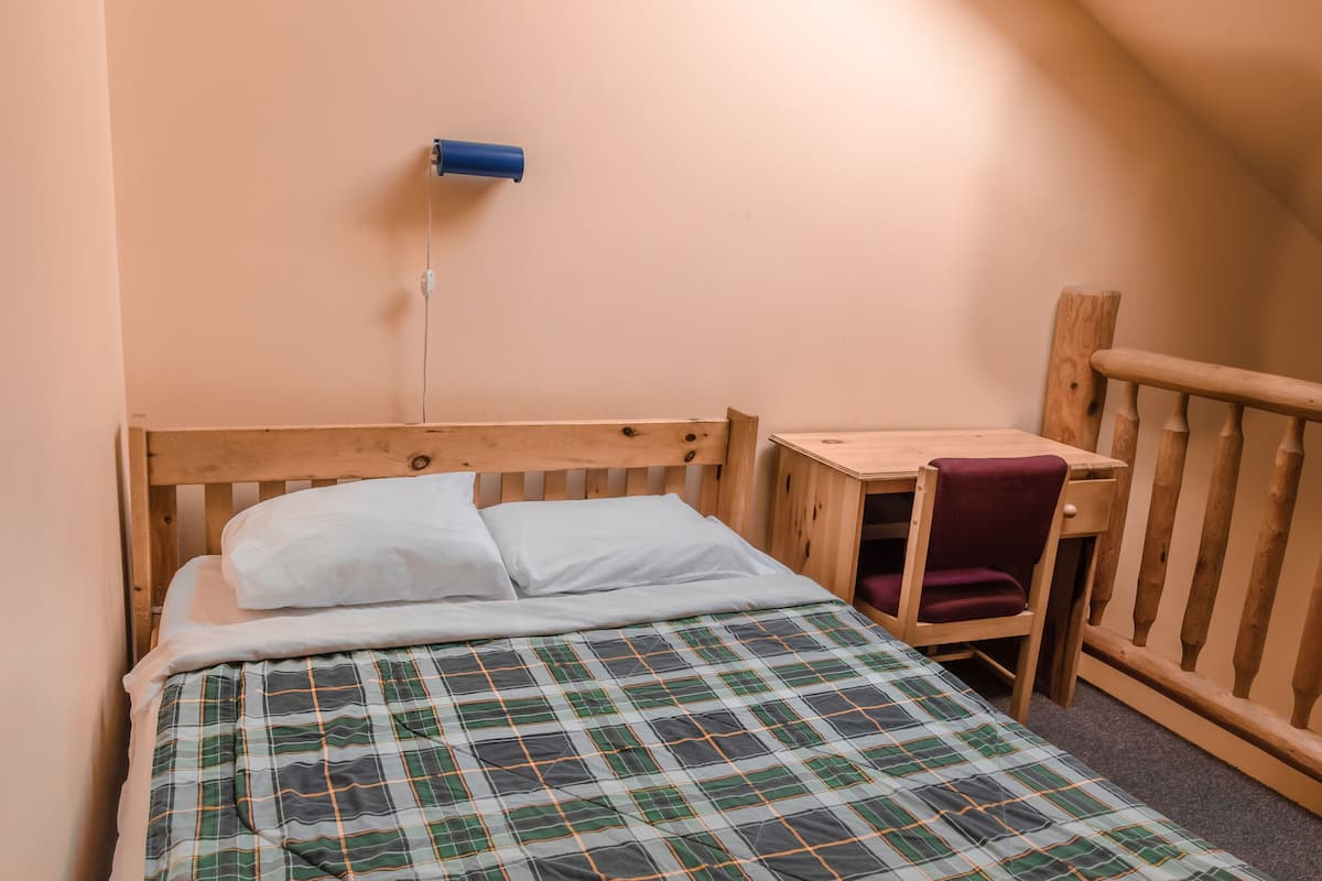 Un lit simple avec une couverture à carreaux, une tête de lit en bois, un petit bureau en bois avec une chaise et une lampe bleue fixée au mur.
