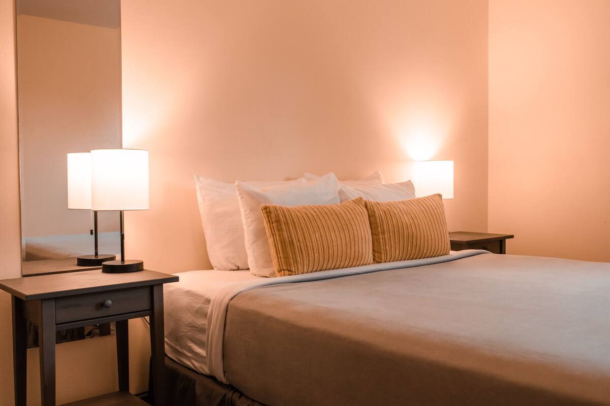 Un lit bien fait, avec des oreillers blancs et bruns, une table de chevet avec une lampe et un miroir accroché au mur.
