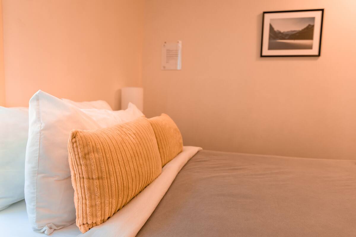 Un lit bien fait, avec des oreillers blancs et beige, un tableau encadré au mur et un petit panneau.