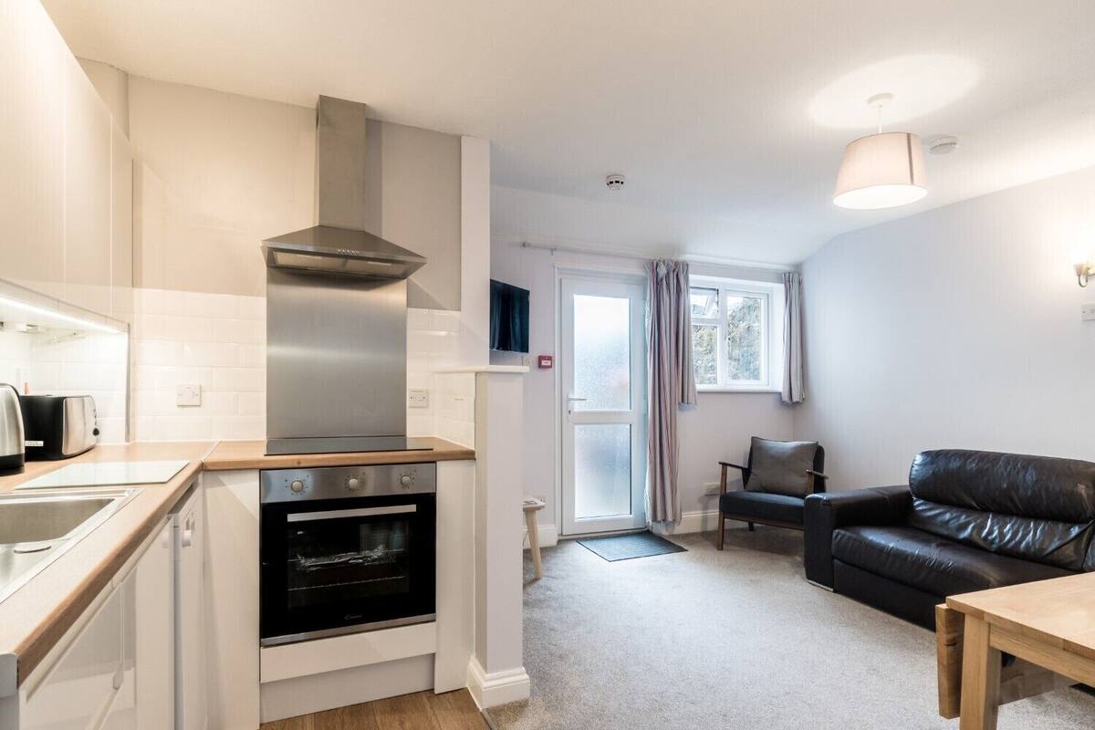 A compact kitchen with modern appliances, a dining area with a black leather sofa, and a window with curtains.