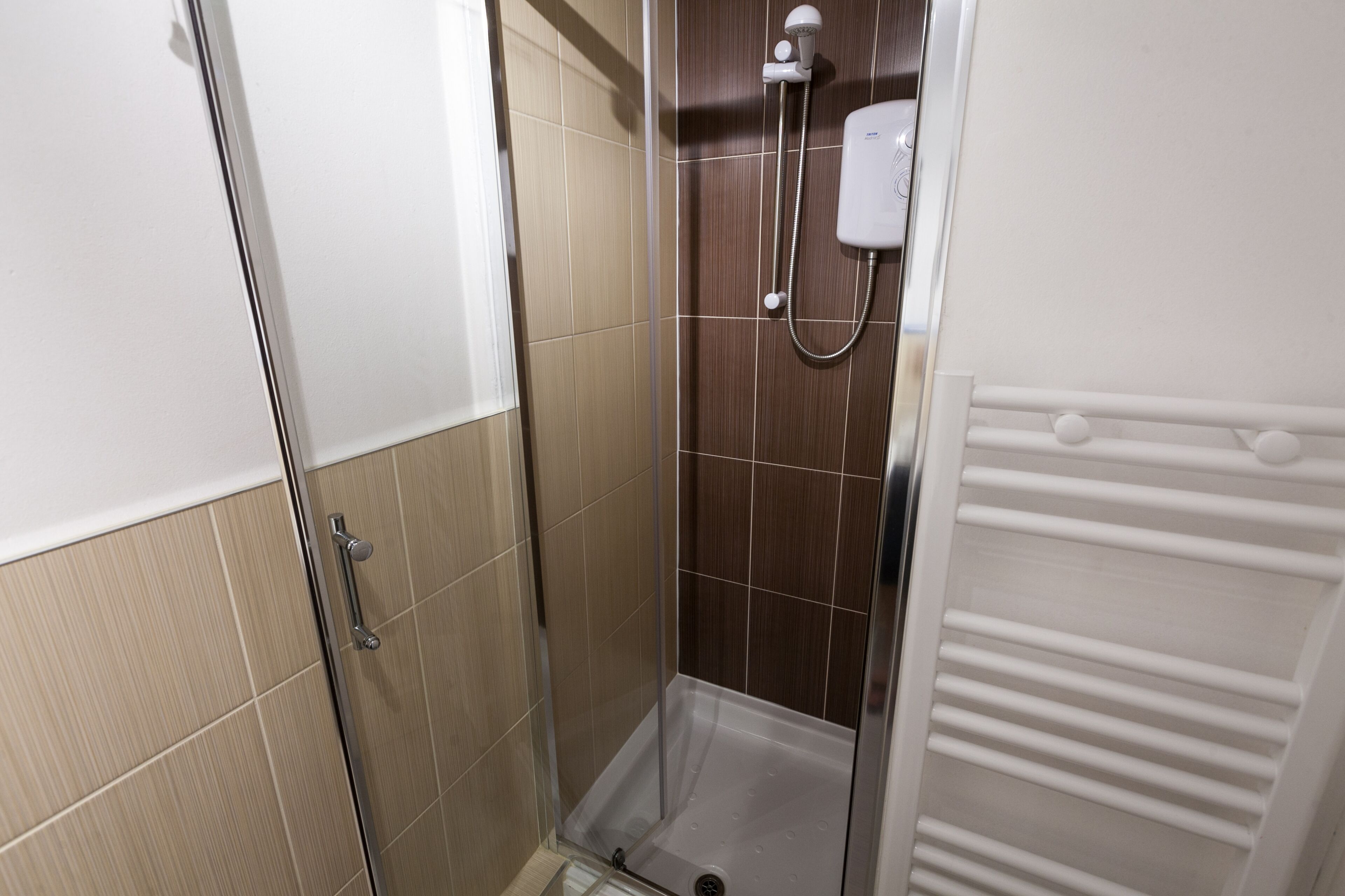 Área de duche com porta de vidro, chuveiro manual e toalheiro branco.
