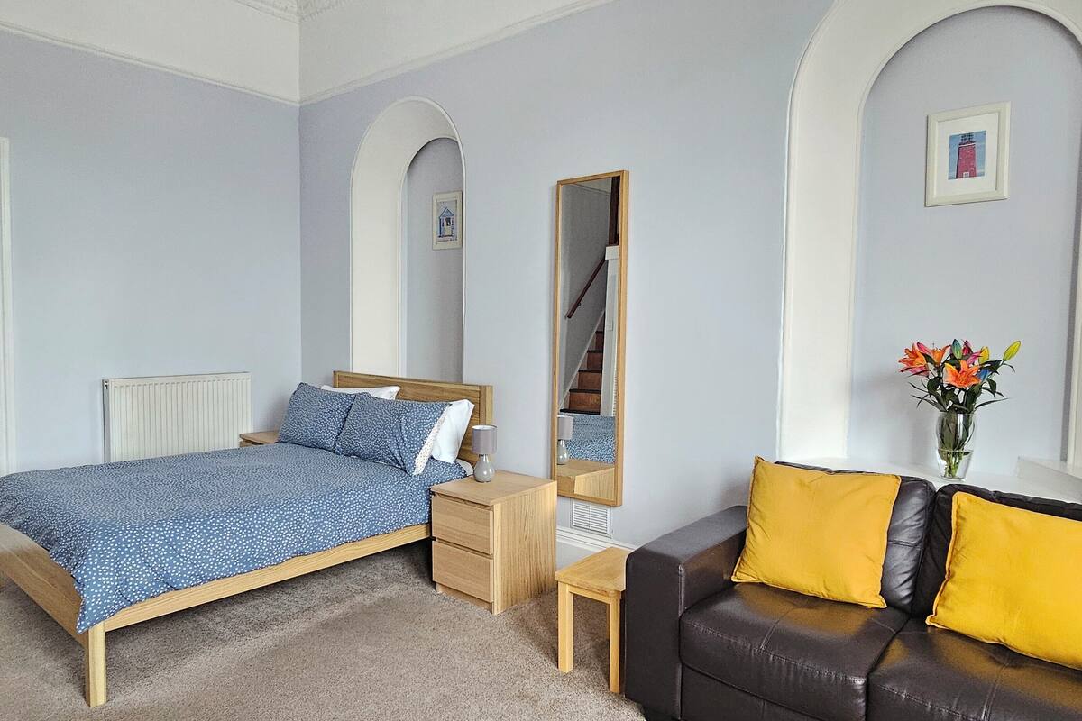 A bedroom with a bed, bedside table, mirror, and a sofa with cushions.