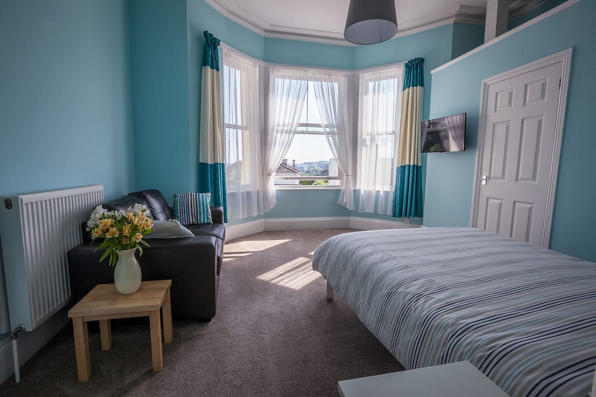 A bedroom with a bed, a sofa, a small table with a vase, and a window with curtains.