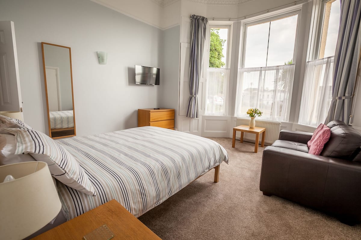 A bedroom with a bed, a dresser, a TV, a window with curtains, a sofa, and a small table with a vase.