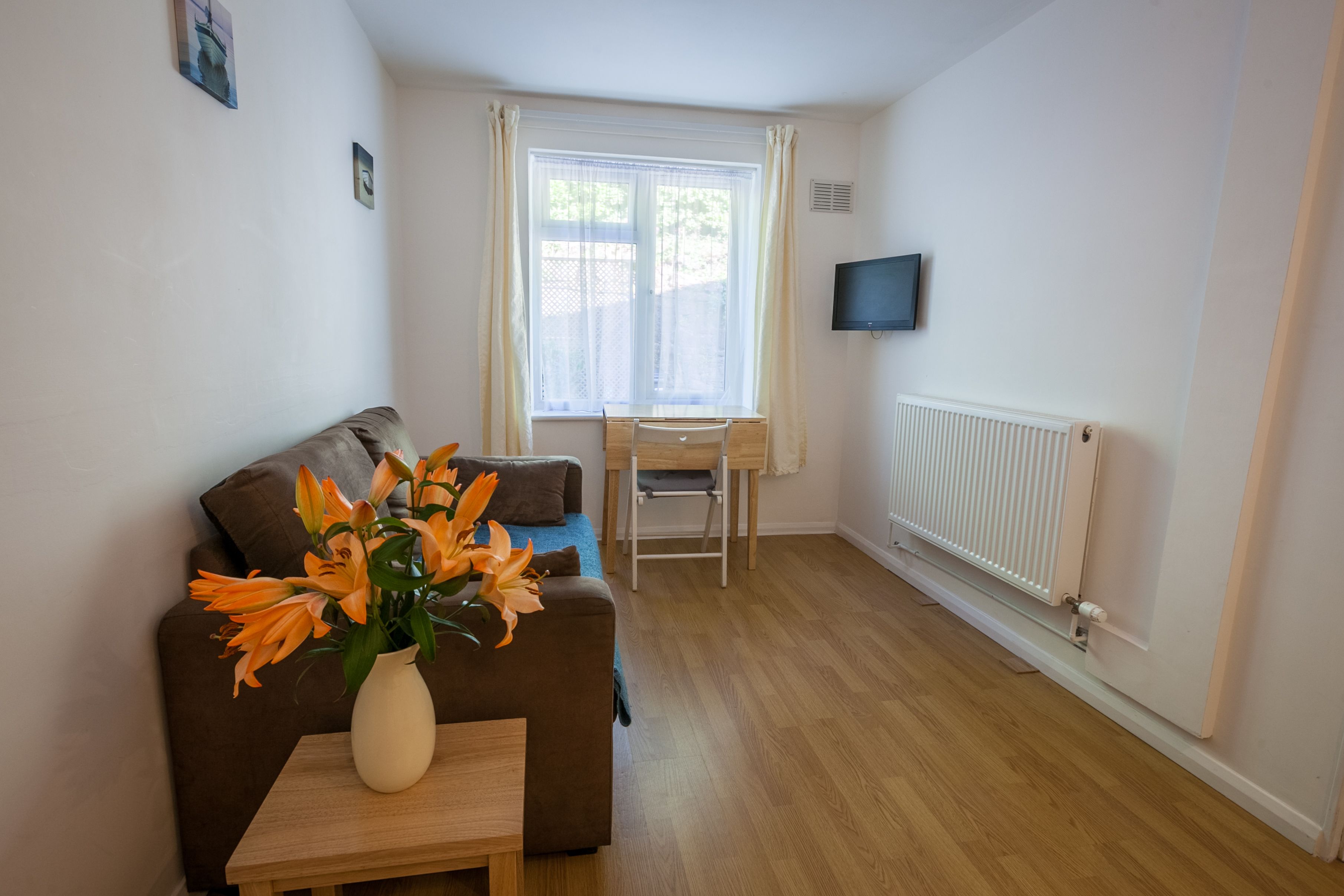 A furnished room with a sofa, a small table with a vase of flowers, a desk with a chair, a radiator, and a TV mounted on the wall.