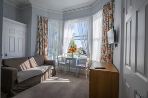 Quarto com sofá, pequena mesa com cadeiras, escrivaninha com cadeira e televisão na parede.
