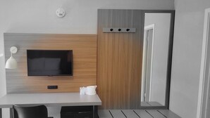 Una habitación moderna con un televisor de pantalla plana montado en un panel de madera, un escritorio blanco con una silla y una lámpara blanca pequeña.