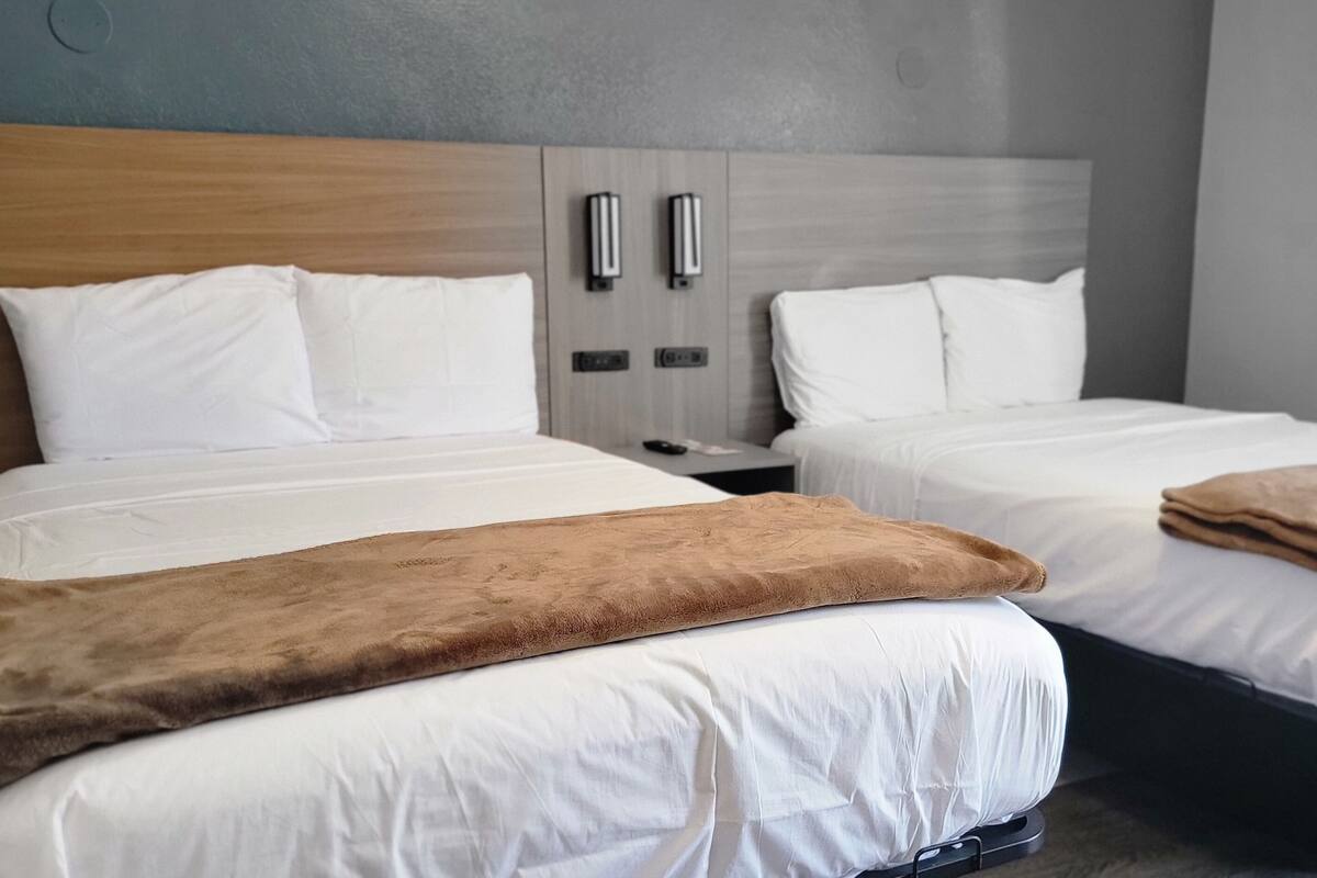 A hotel room with two beds, a wooden headboard, bedside tables, and a window with blinds.
