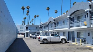 Un estacionamiento con varios vehículos estacionados, incluyendo una camioneta blanca, frente a un edificio con múltiples balcones y palmeras al fondo.