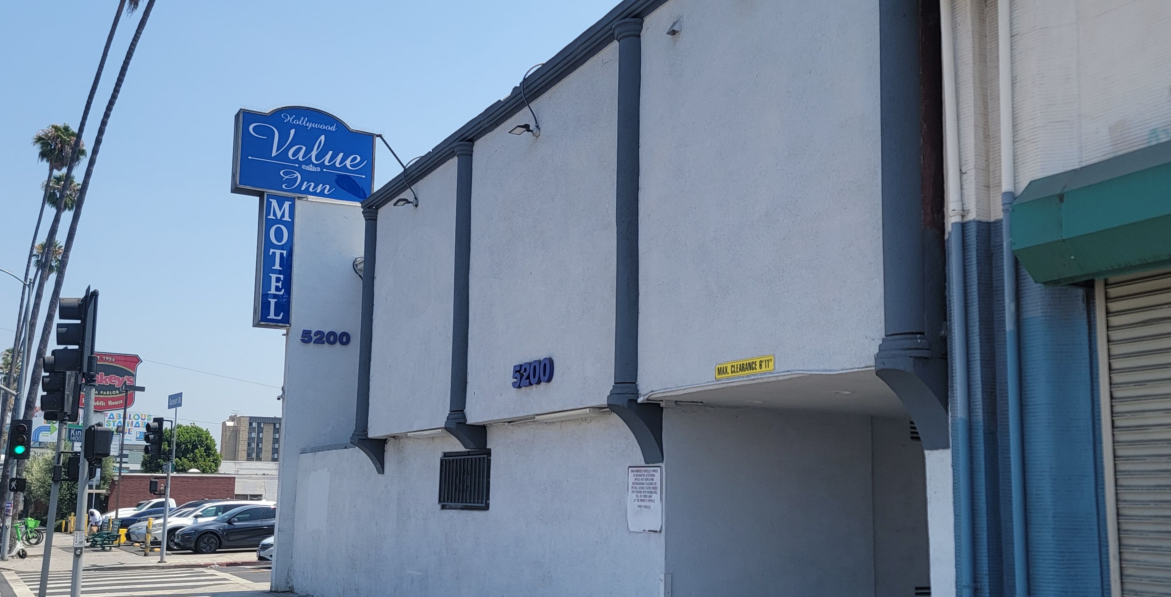 Un edificio con el letrero del Hollywood Value Inn, un motel con el número 5200, y una calle con coches y semáforos.