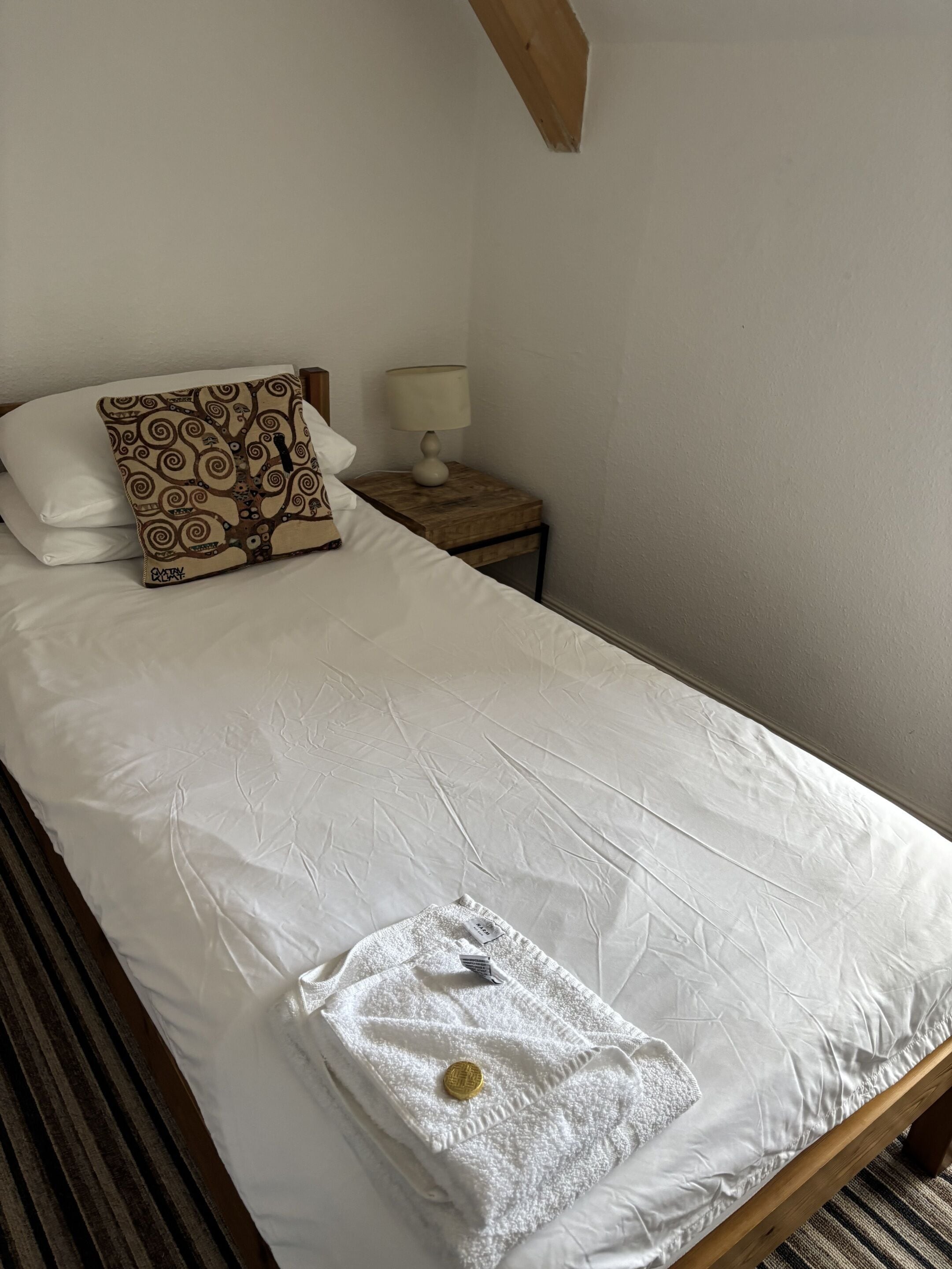 Ein ordentlich bezogenes Bett mit weißer Daunendecke, einem gemusterten Kopfkissen und einem gefalteten weißen Handtuch mit Goldemblem auf einem Streifen Teppich.