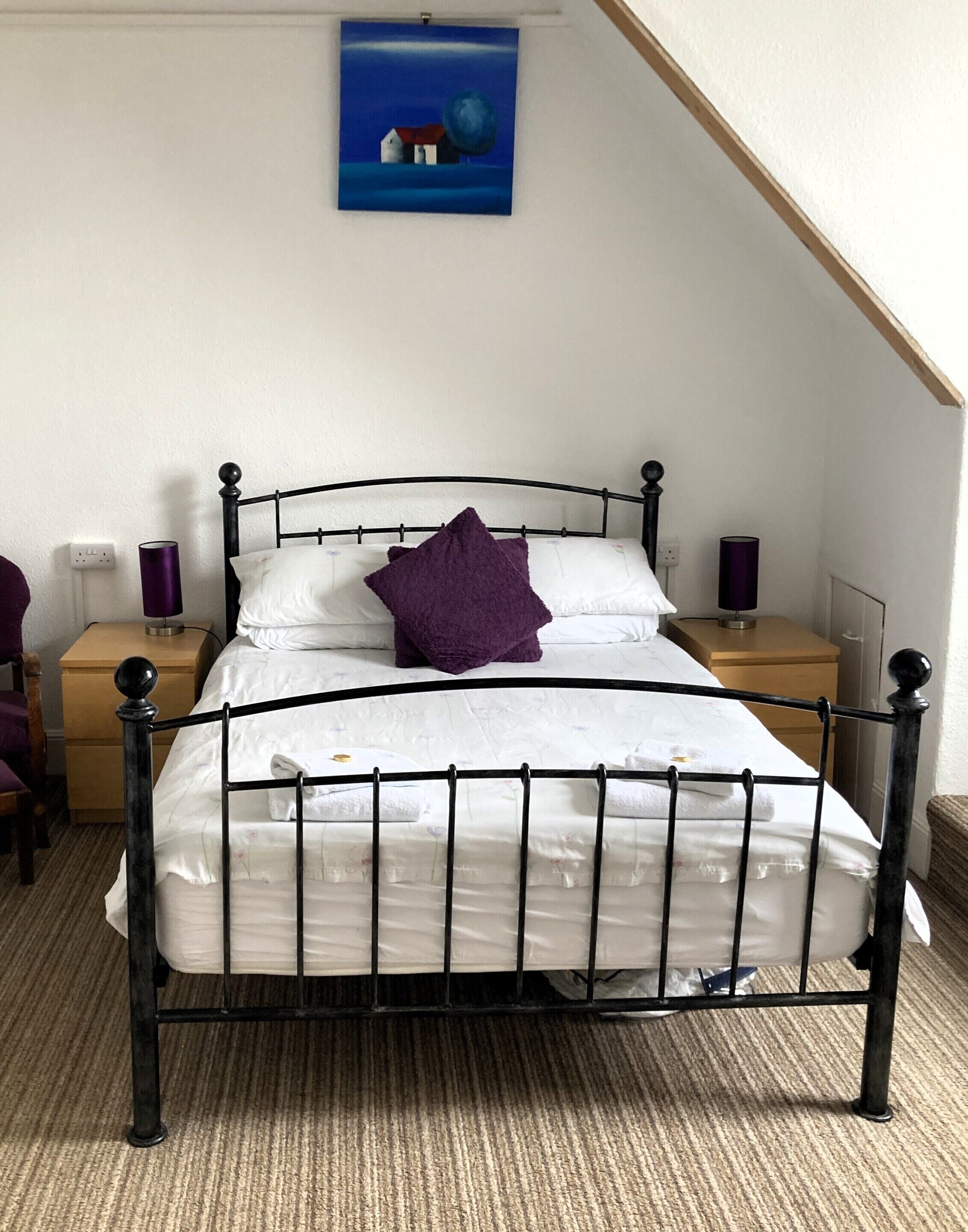 A neatly made bed with a black metal frame, white bedding, and a purple pillow. There are two bedside tables with lamps, a wooden nightstand, and a painting on the wall.