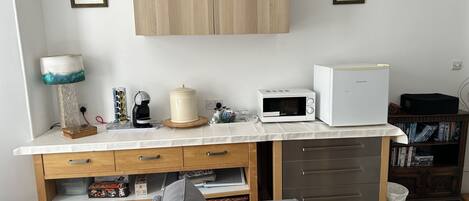 A compact kitchenette with a microwave, toaster, and coffee maker. There are two framed pictures on the wall, a lamp, and a small cabinet below the counter.