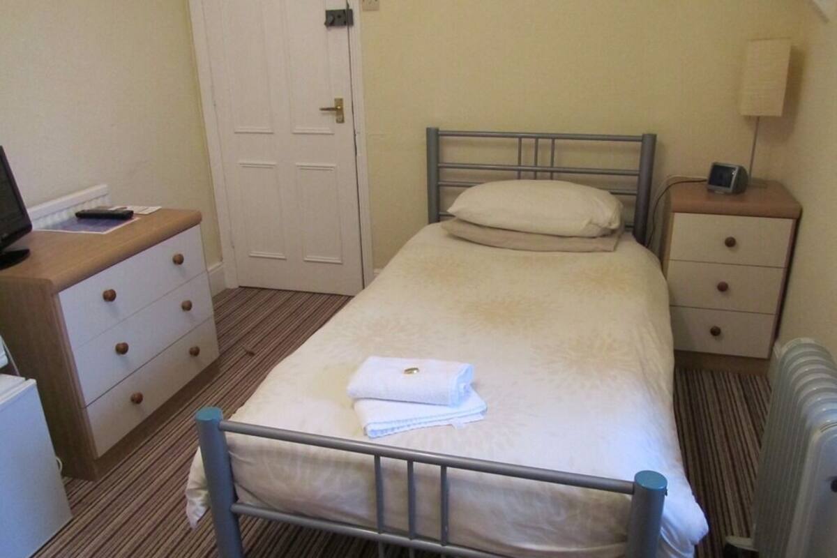 Una habitación pequeña y limpia con una cama individual, una mesita de noche con lámpara, un aparador y una puerta.