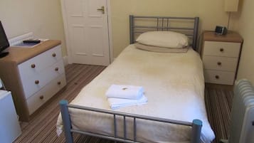 A small, clean room with a single bed, a bedside table with a lamp, a dresser, and a door.