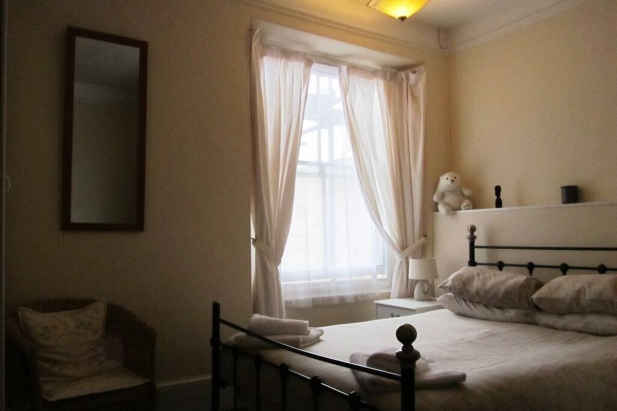 Un dormitorio con cama, una silla, una mesita de noche, una lámpara y una ventana con cortinas.