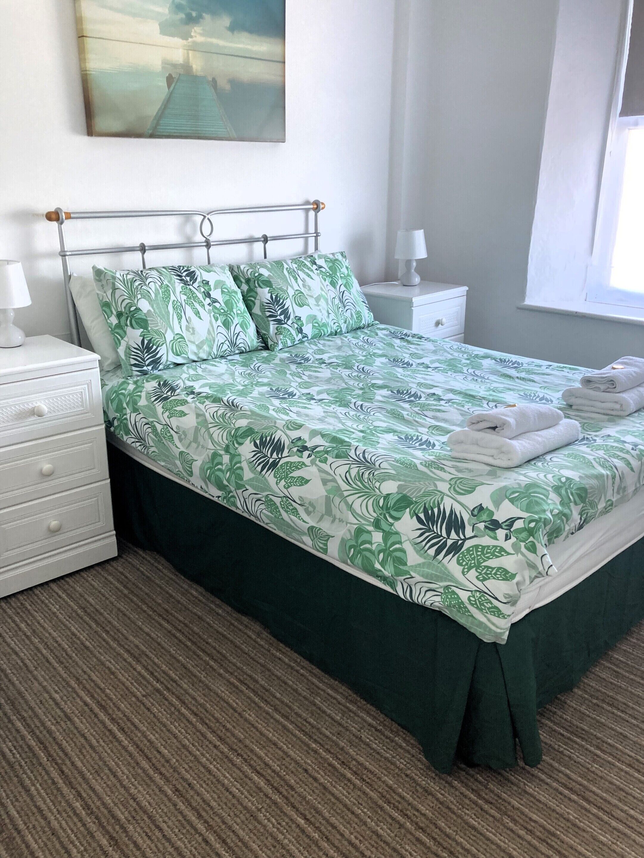 Een keurig opgemaakt bed met een spreideken met een groen-wit bladmotief, witte kussens en een metalen hoofdbord. Er staat een witte nachtkastje met een lamp en een lade, en een raam met witte gordijnen.