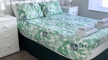 A neatly made bed with a green and white leaf pattern bedspread, white pillows, and a metal headboard. There is a white nightstand with a lamp and a drawer, and a window with white curtains.