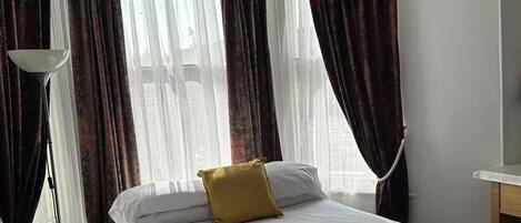 Habitación de hotel con una cama bien hecha, una lámpara de pie y vistas a través de cortinas translúcidas.