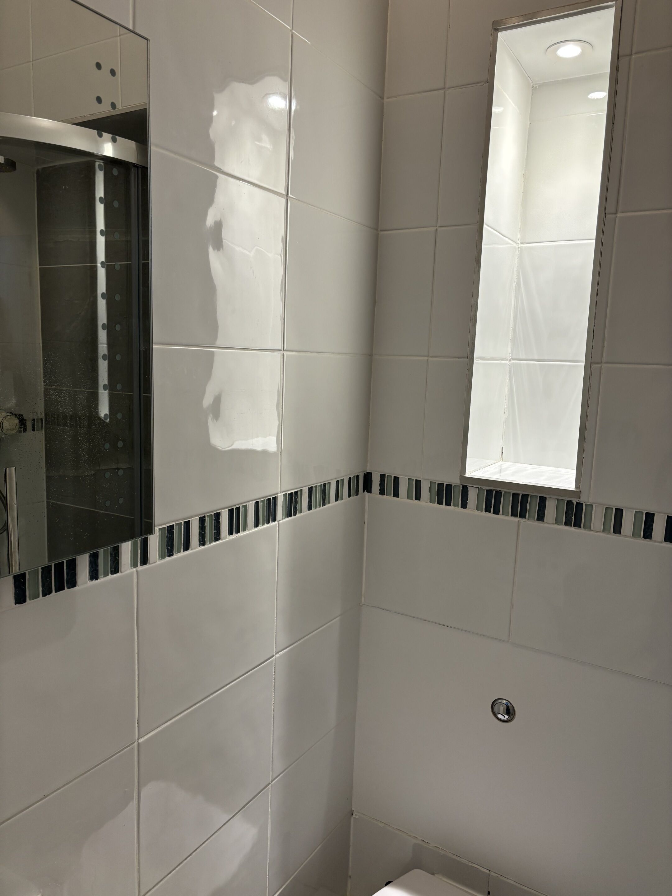 A bathroom with white tiles, a mirror, a window, and a shower.
