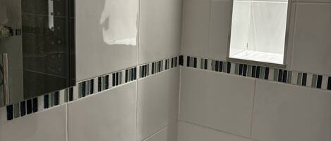 A bathroom with white tiles, a mirror, a window, and a shower.