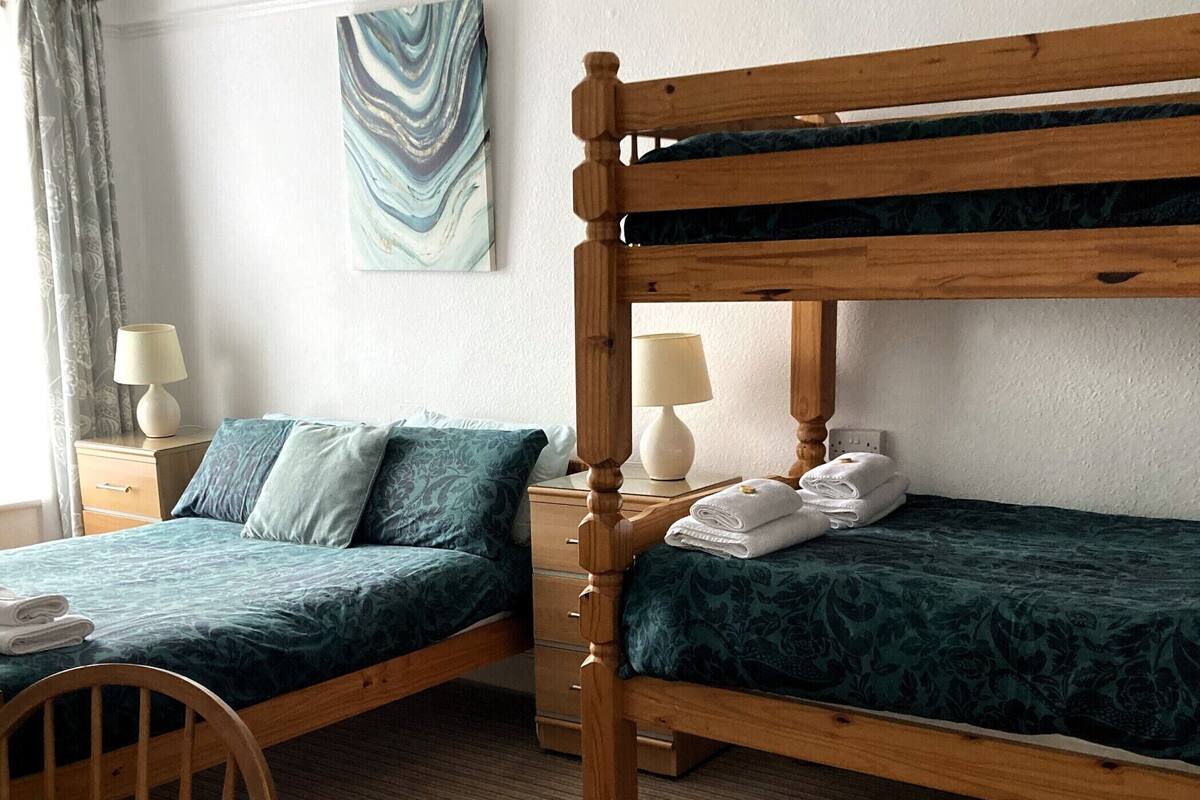 Una habitación de hotel con una litera, una cama individual, una mesa de madera y una silla. Hay una lámpara en la mesita de noche y un cuadro en la pared.