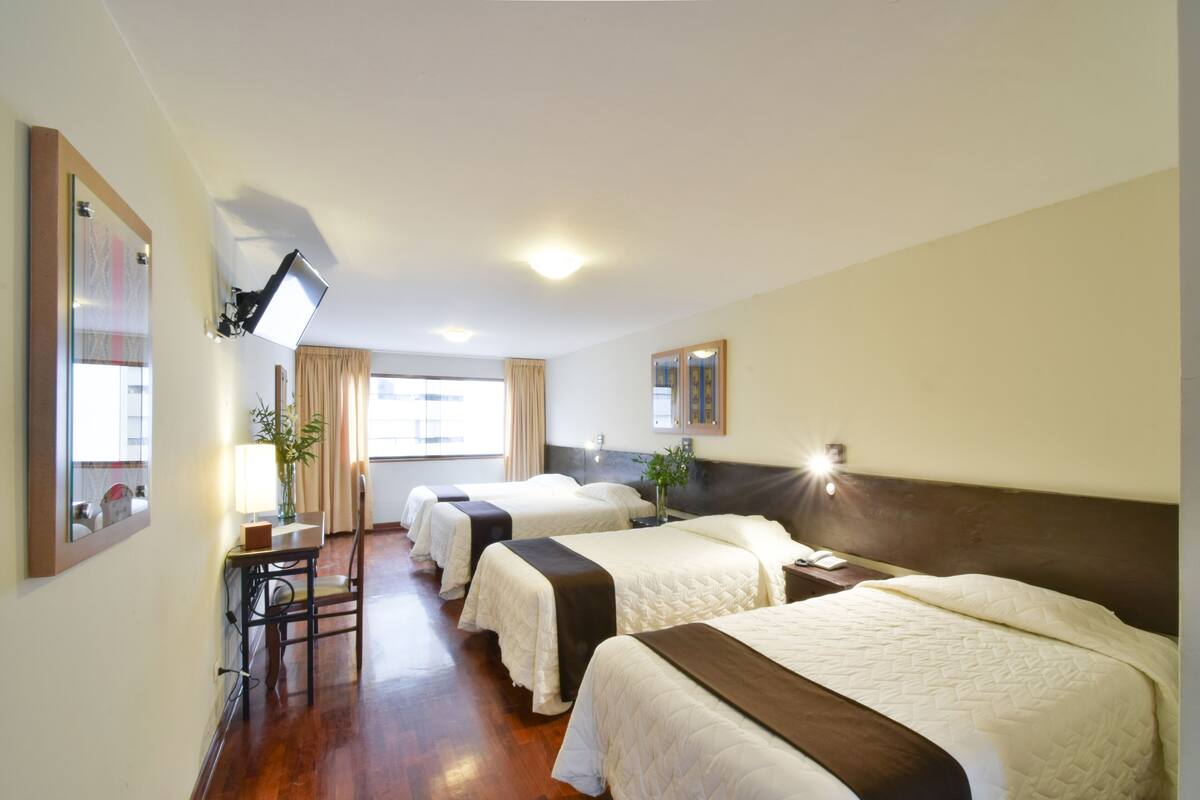 Quarto de hotel com três camas, uma escrivaninha com luminária, uma TV fixada na parede e uma janela com cortinas.