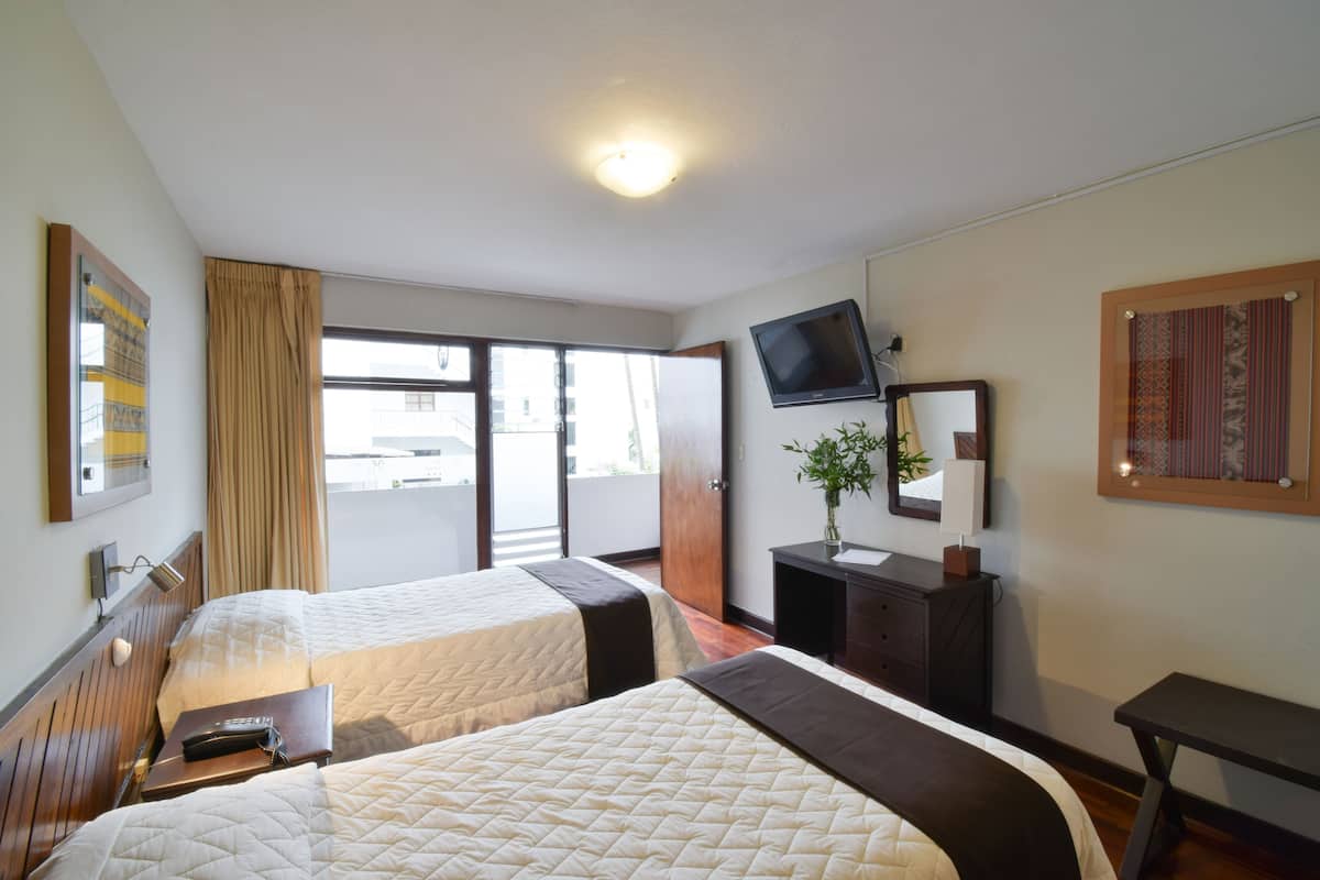 Quarto de hotel com duas camas, televisão na parede, secretária com uma planta e porta de correr para a varanda.