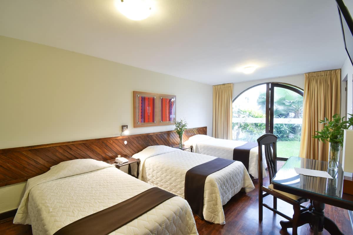 Quarto de hotel com duas camas, cabeceira de madeira, mesa de jantar com cadeiras, um vaso com planta e uma janela com cortinas.