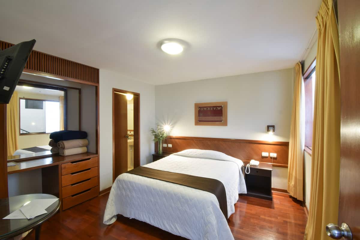 Quarto de hotel com cama, escrivaninha, espelho, mesa de cabeceira e televisão na parede.