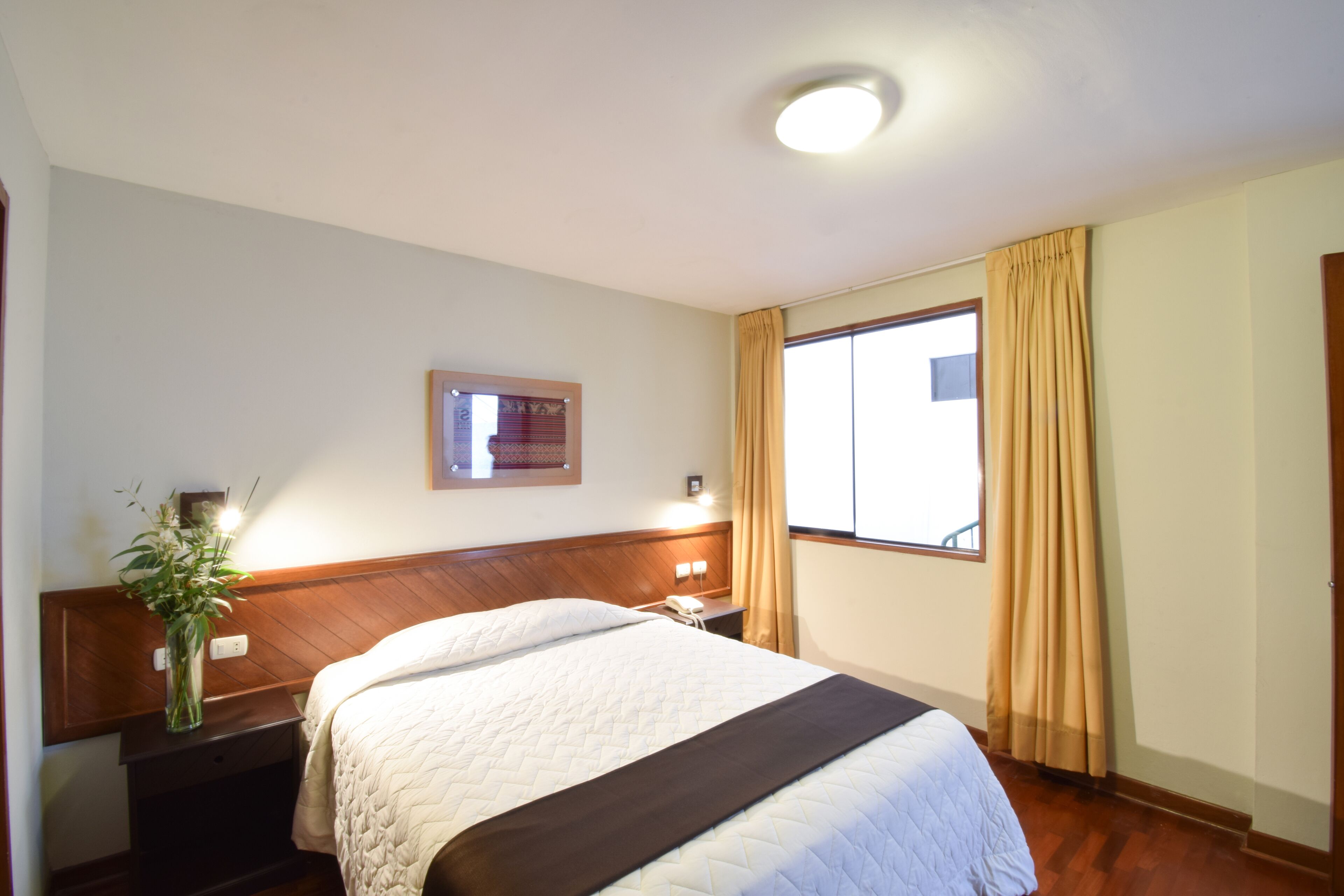 Hotelkamer met een bed, nachtkastje, lamp, raam met gordijnen en een aan de muur hangend schilderij.