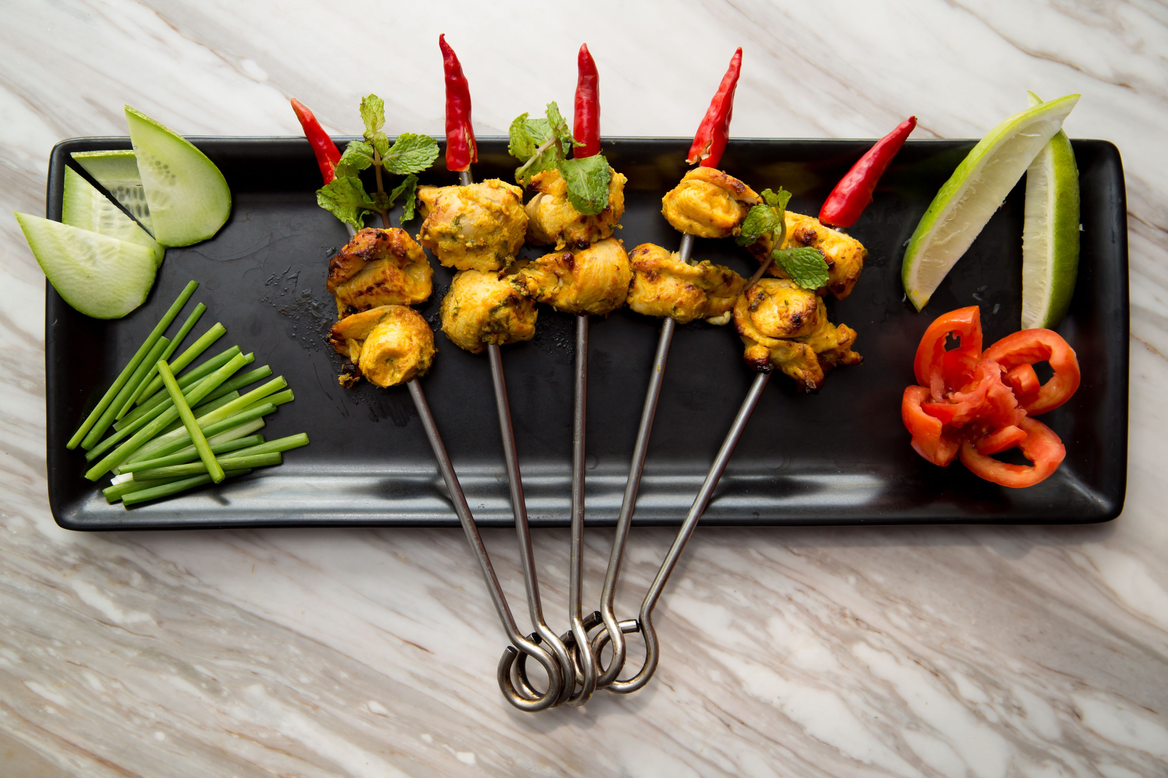 Un plato rectangular negro con brochetas de pollo a la parrilla, rodajas de pepino, cebolletas, chiles rojos y gajos de lima.