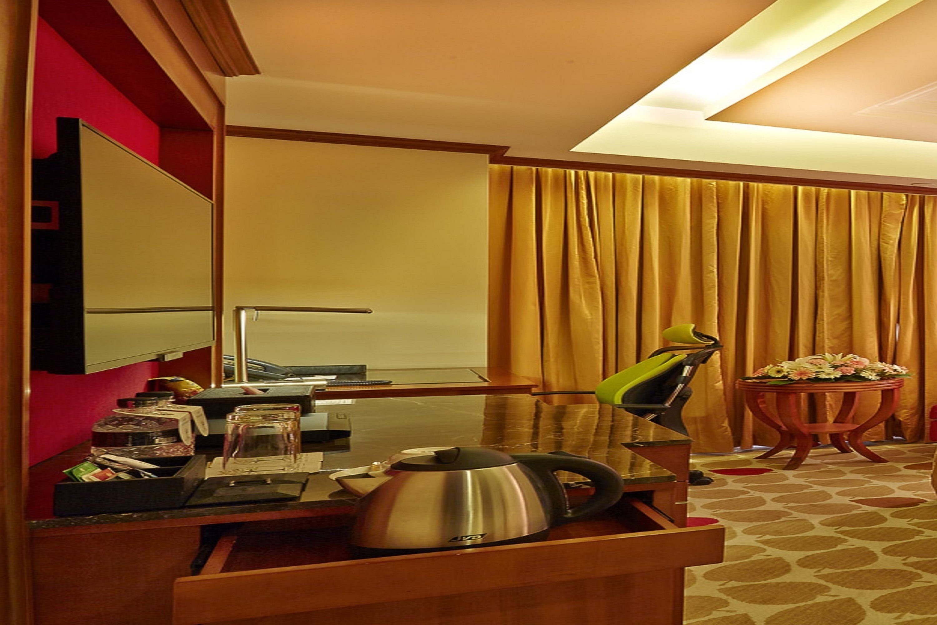 A hotel room with a desk, a kettle, a chair, a small table with flowers, and a large window with curtains.