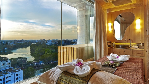 A hotel room with a large window offering a city view, a wooden vanity with a mirror, and a bed with towels and a flower. - Six Seasons Hotel (Dhaka)