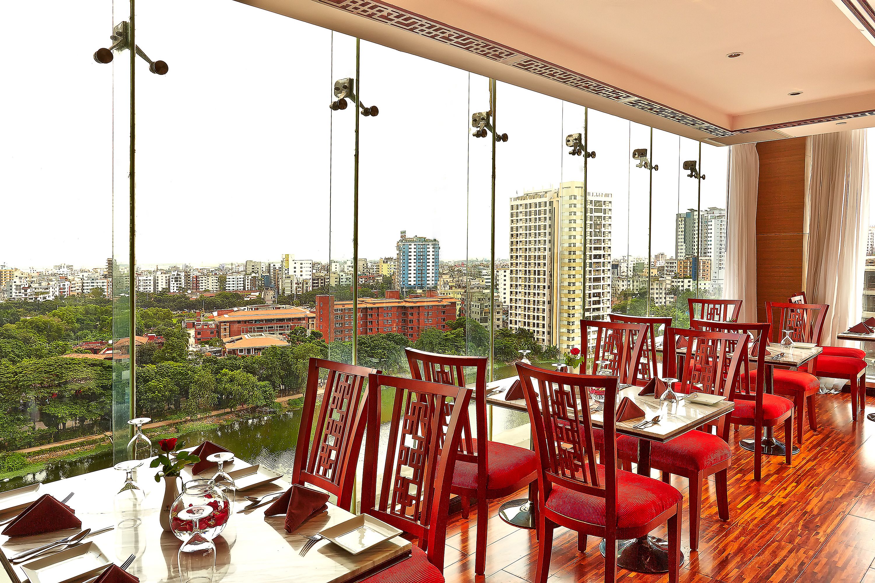 A dining area with red chairs and tables, large windows offering a city view, and a high ceiling with hanging lights.