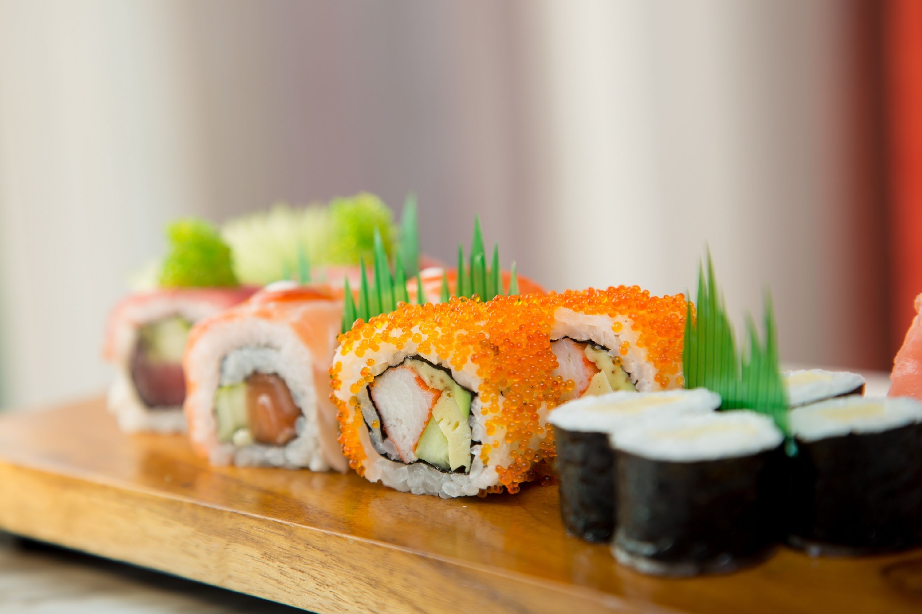 A wooden board with various sushi rolls, including nigiri and maki, garnished with green leaves.