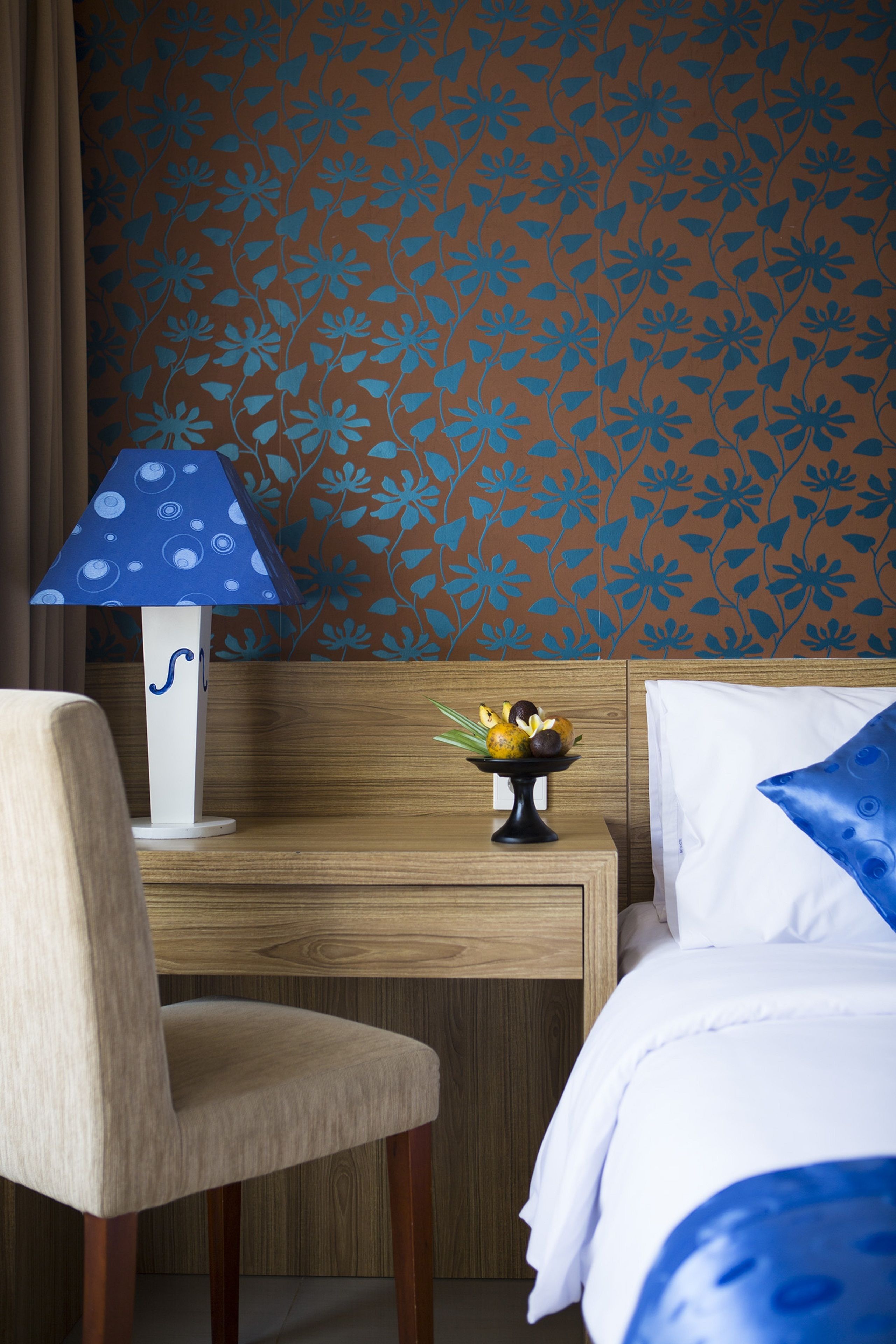 Habitación de hotel con cabecera de madera, lámpara azul estampada, frutero pequeño y cama con ropa de cama blanca y azul.