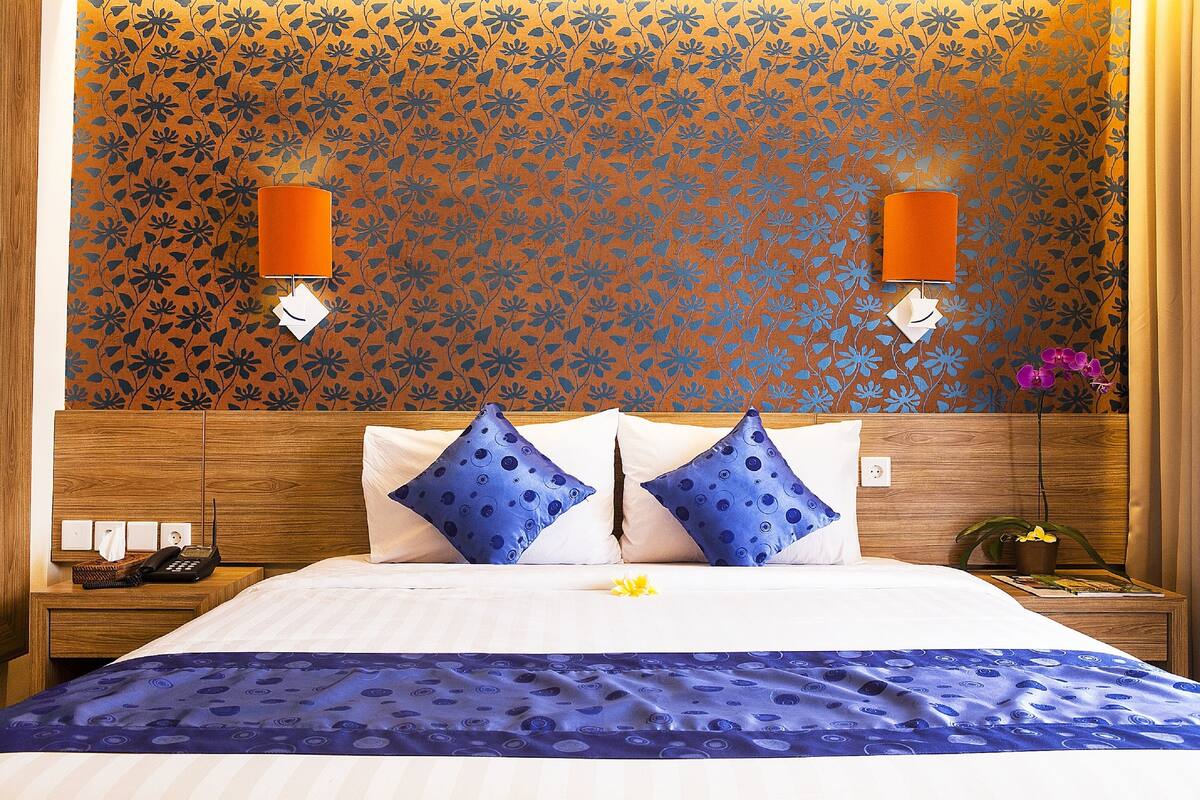 Une chambre d’hôtel avec un papier peint à motifs floraux, un lit avec une literie bleue et blanche, deux oreillers bleus et une table de chevet avec un téléphone et une lampe.